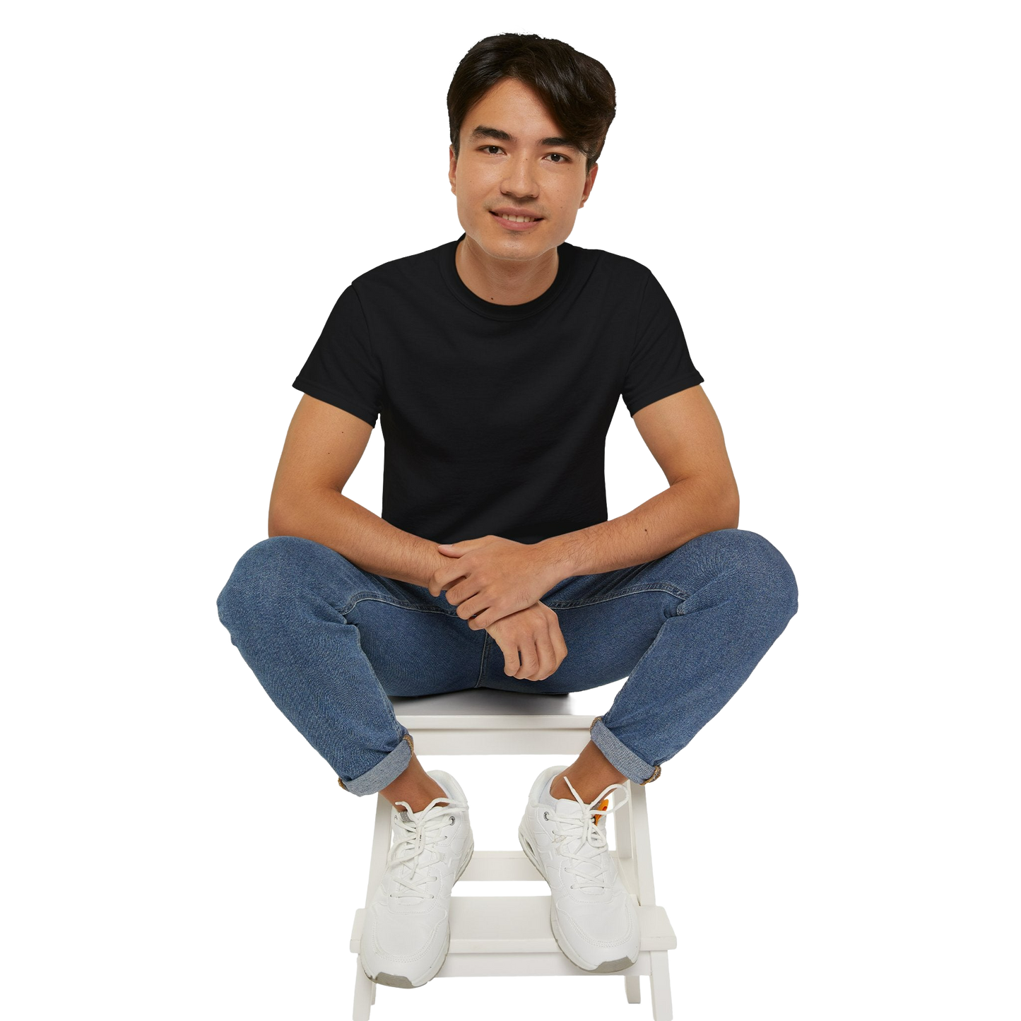 A young man wearing a black t-shirt and blue jeans is sitting on a white stool, with his hands crossed and a smile on his face.