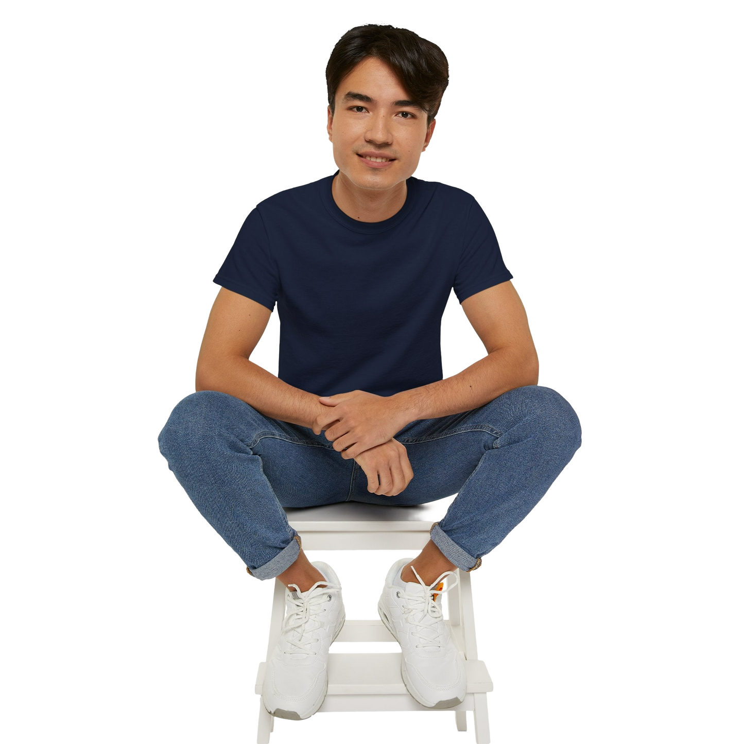 A young man wearing a navy blue t-shirt and blue jeans is sitting on a white stool, with his hands crossed and a smile on his face.