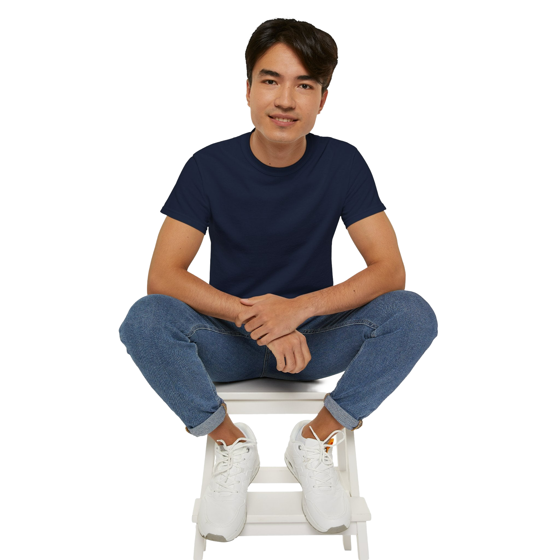 A young man wearing a navy blue t-shirt and blue jeans is sitting on a white stool, with his hands crossed and a smile on his face.