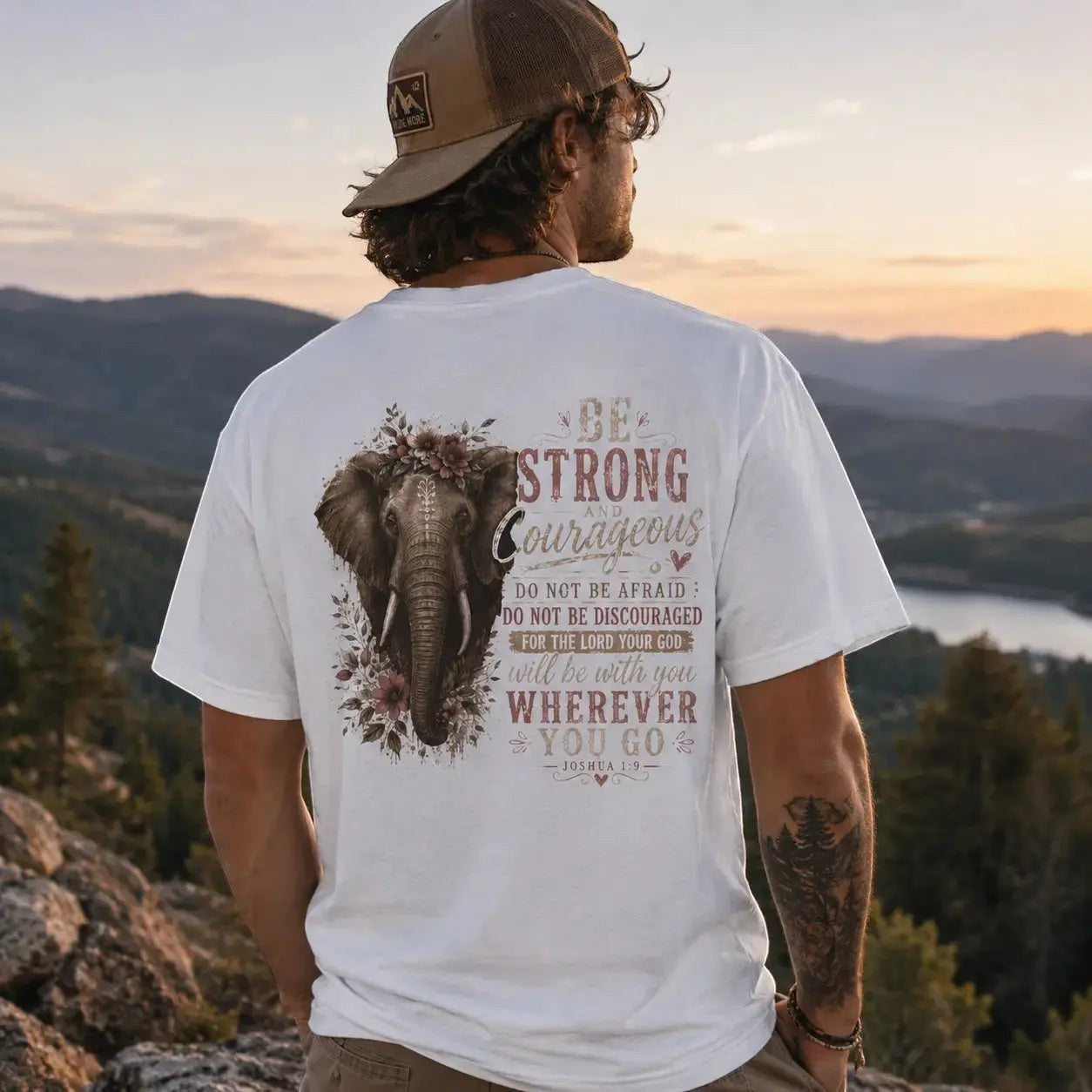 Man wearing a Just Chilling Shop white graphic t-shirt with an elephant and floral design at a mountain overlook.